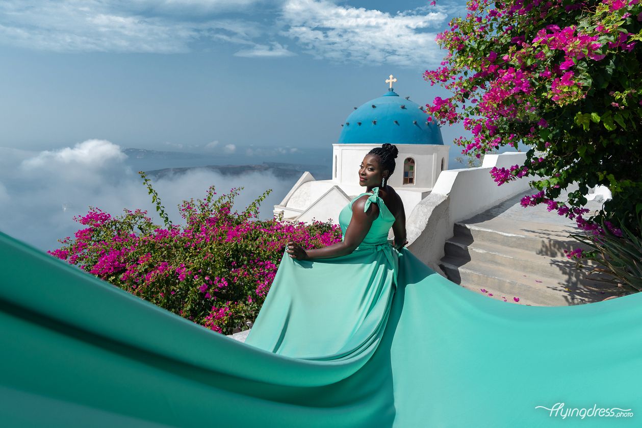 A stunning flying dress photoshoot in Santorini, featuring a woman in a flowing turquoise gown against iconic blue-domed churches and vibrant bougainvillea. Elegant, dreamy, and perfect for capturing the magic of the island.